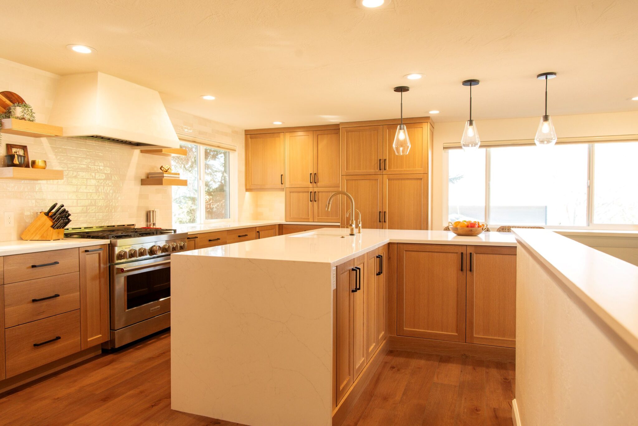 Modern kitchen remodel featuring a waterfall-edge white quartz island with built-in sink and faucet, light wood cabinetry with black handles, stainless steel appliances, white subway tile backsplash, floating wood shelves, and pendant lighting. Natural light streams through large windows, highlighting the warm wood flooring and clean, contemporary design.